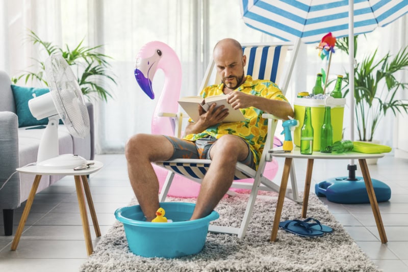 3 Signs It’s Time for a New AC. Image is a photograph of a man spending his summer vacations at home, he is sitting on a deckchair in the living room and reading a book.