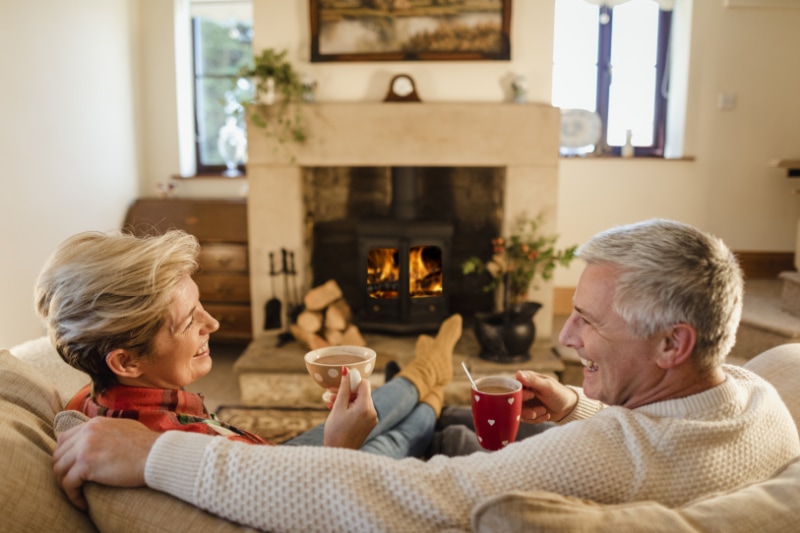 Mature couple sat infront of a fire drinking hot chocalate from mugs.