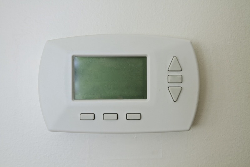 Why Is My Thermostat Blank? Image is a photograph of a bland thermostat mounted on a white wall.