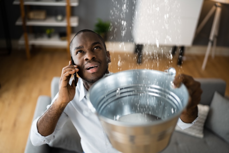 How Can I Avoid a Plumbing Emergency? Man on phone calling plumber catching leaking water from ceiling in a bucket.